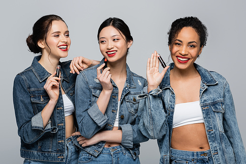 Smiling multiethnic women in denim jackets holding lipsticks isolated on grey