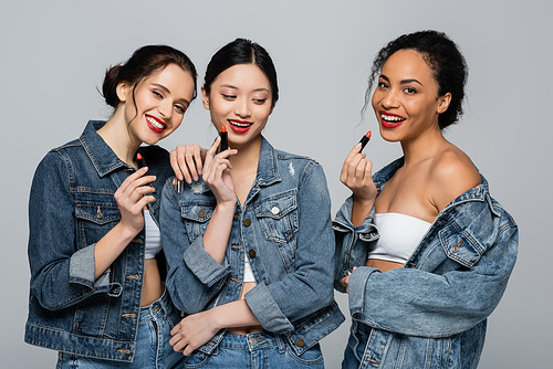Pretty multiethnic women in denim jackets smiling while holding lipsticks isolated on grey