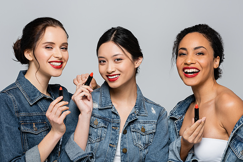 Smiling interracial friends holding red lipsticks isolated on grey