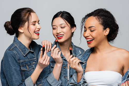 Pretty interracial women in denim jackets holding lipsticks isolated on grey