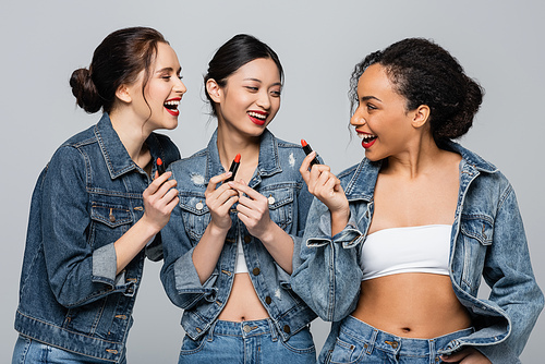 Happy multiethnic women in denim jackets holding red lipsticks isolated on grey