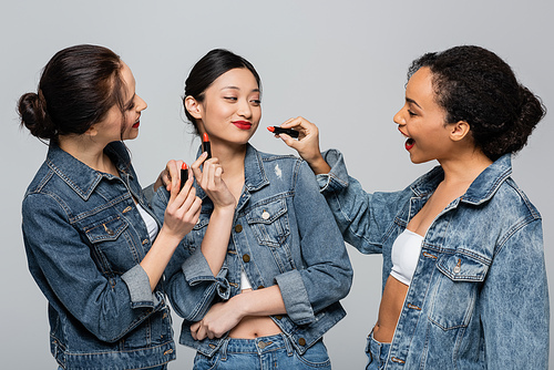 Smiling multiethnic women in denim jackets holding red lipsticks near asian friend isolated on grey