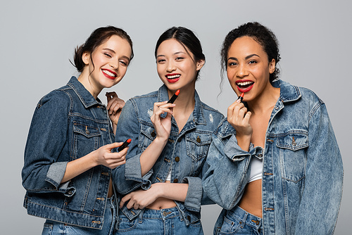 Pretty multiethnic women in denim jackets holding red lipsticks and  isolated on grey