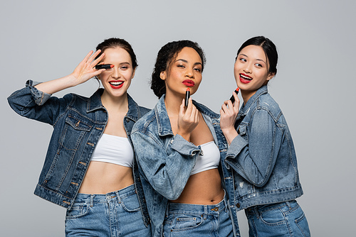 Young interracial women in denim jackets holding red lipsticks isolated on grey