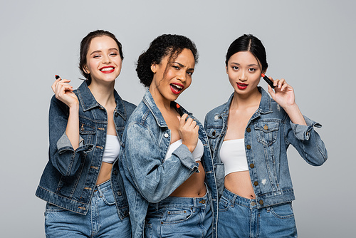 African american woman holding red lipsticks near interracial friends in denim jackets isolated on grey