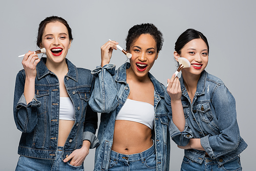Happy multicultural women in denim jackets holding cosmetic brushes isolated on grey