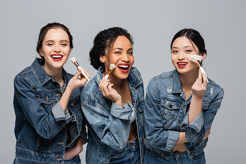 Happy multicultural women with red lipsticks holding cosmetic brushes isolated on grey