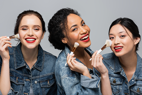 Cheerful interracial women with red lips holding cosmetic brushes near faces isolated on grey