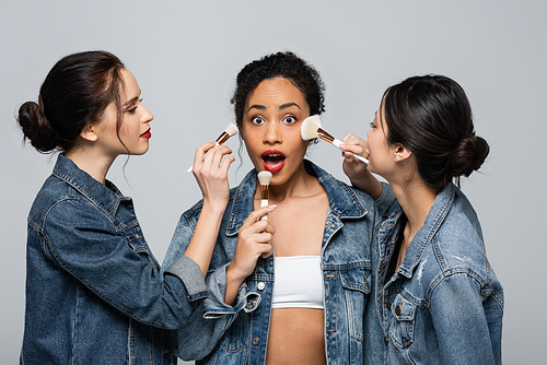 Young interracial women in denim jackets holding cosmetic brushes near shocked african american friend isolated on grey