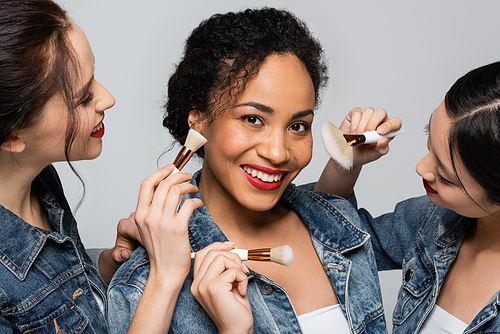 Smiling african american woman in denim jacket standing near interracial friends with cosmetic brushes isolated on grey