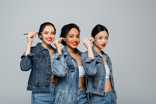 Cheerful interracial women in denim jackets holding cosmetic brushes near eyes isolated on grey