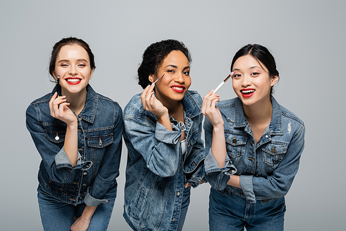 Smiling multiethnic women in denim jackets holding cosmetic brushes and  isolated on grey