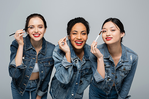 Happy interracial women with red lips holding cosmetic brushes isolated on grey