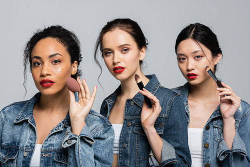 Young multiethnic women in denim jackets holding cosmetic brushes and beauty blender isolated on grey