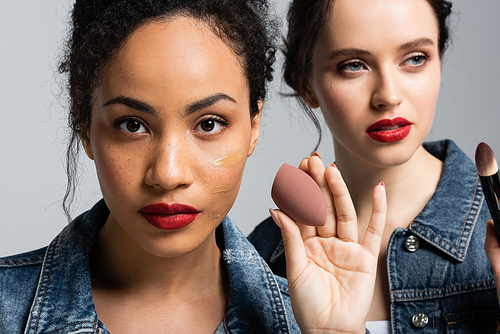 African american woman with face foundation holding beauty blender near friend with cosmetic brush isolated on grey