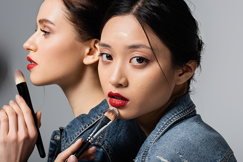 Asian woman with red lips and face foundation holding cosmetic brush near friend on grey background