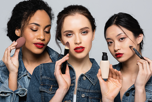 Young woman holding face foundation and cosmetic brush near interracial friends isolated on grey