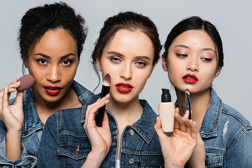 Interracial women with red lips holding cosmetic brushes and face foundation isolated on grey
