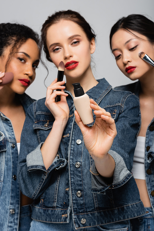 Face foundation in hand of blurred woman near interracial friends with cosmetic brushes isolated on grey