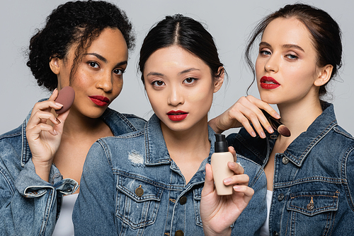Asian woman holding face foundation near interracial friends with beauty blender and cosmetic brush isolated on grey