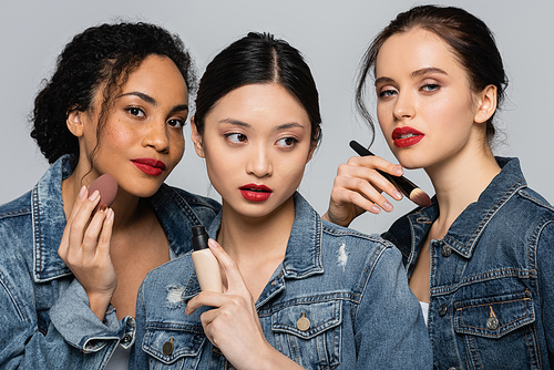 Interracial young women holding cosmetic brush, face foundation and beauty blender isolated on grey