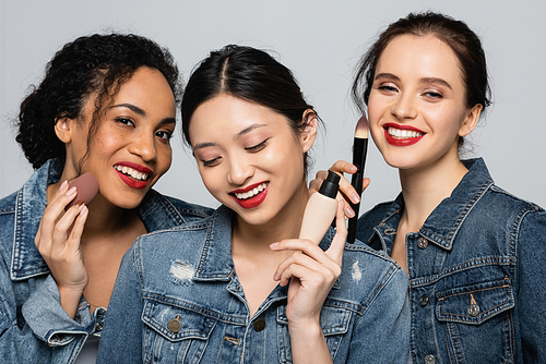 Smiling interracial women holding face foundation, cosmetic brush and beauty blender isolated on grey