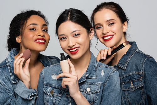 Smiling asian woman holding face foundation near interracial friends with red lips isolated on grey