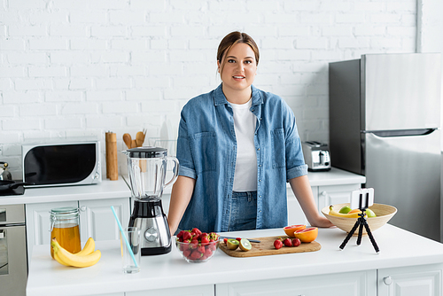 Cheerful body positive woman  near fruits, honey and smartphone in kitchen
