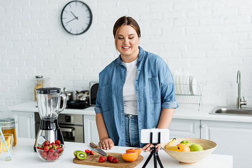 Woman with overweight looking at smartphone near fresh fruits and blender in kitchen