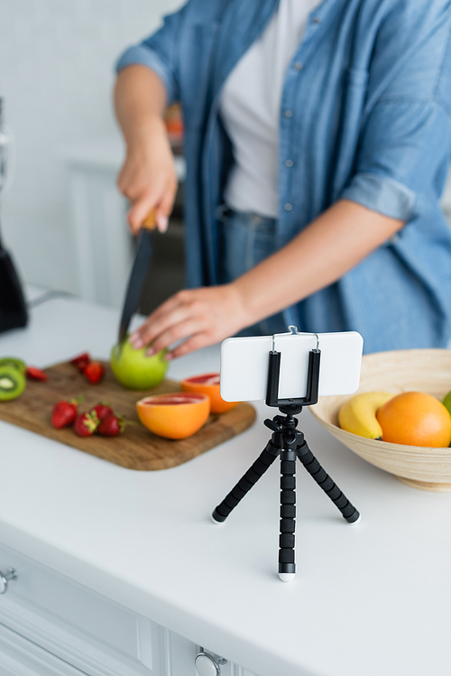 Cropped view of smartphone near blurred woman cutting fruits in kitchen
