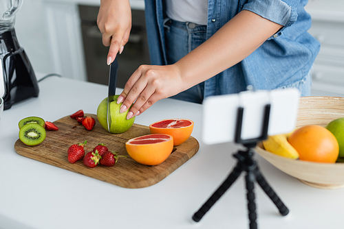 Cropped view of young woman cutting fruits near smartphone on tripod in kitchen