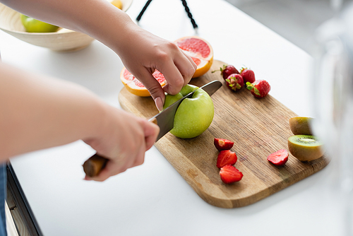 Cropped view of woman cutting apple near strawberries and kiwi in kitchen