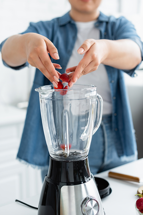 Cropped view of blurred plus size woman putting strawberries in blender in kitchen