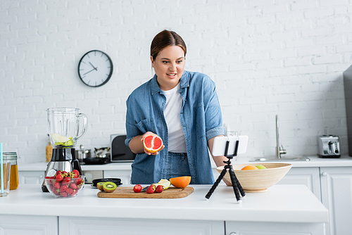 Smiling plus size woman holding grapefruit near fruits and smartphone in kitchen