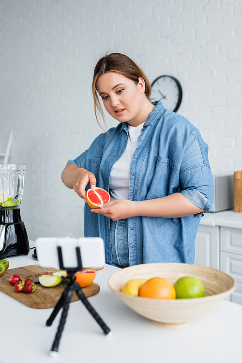 Plus size woman holding grapefruit near blurred smartphone and blender in kitchen