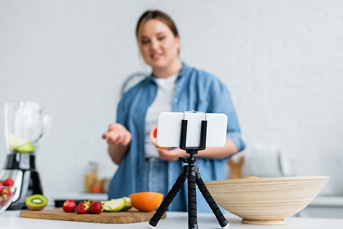 Smartphone on tripod near blurred plus size woman and fruits in kitchen
