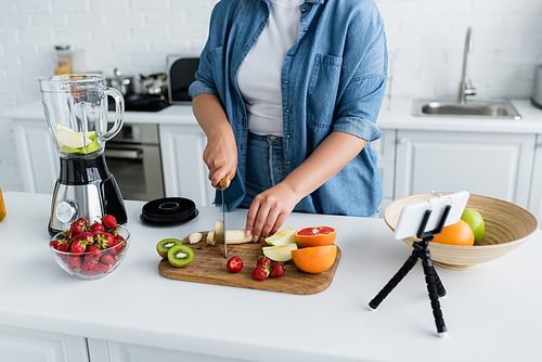 Cropped view of woman with overweight cutting fruits near blender and smartphone