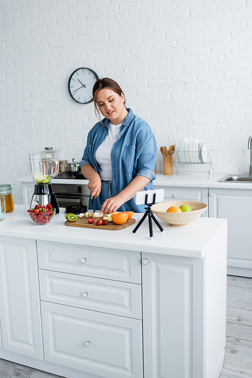 Smiling plus size woman cutting fresh fruits near blender and smartphone in kitchen
