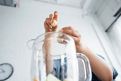 Low angle view of blurred woman holding sliced banana above blender in kitchen