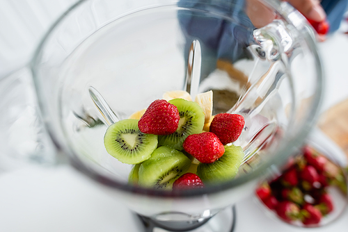 Cropped view of fresh fruits in blender near blurred woman in kitchen