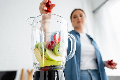 Low angle view of blurred plus size woman pouring fruits in blender in kitchen