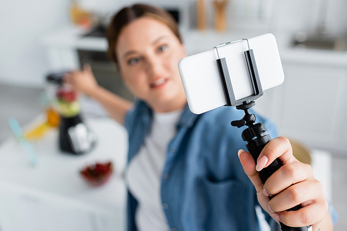 High angle view of smartphone on tripod in hand of blurred plus size woman in kitchen