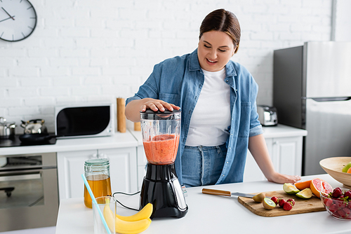 Smiling plus size woman preparing smoothie in blender near honey and fresh fruits in kitchen