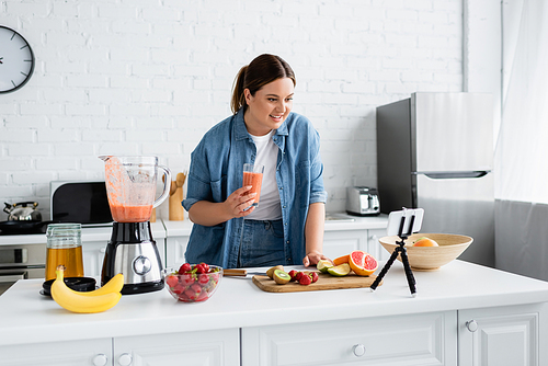 Cheerful plus size woman holding glass of smoothie near fruits and smartphone in kitchen