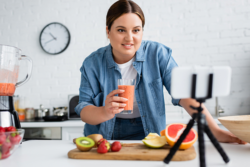 Positive plus size woman holding glass of smoothie near blurred cellphone on tripod in kitchen