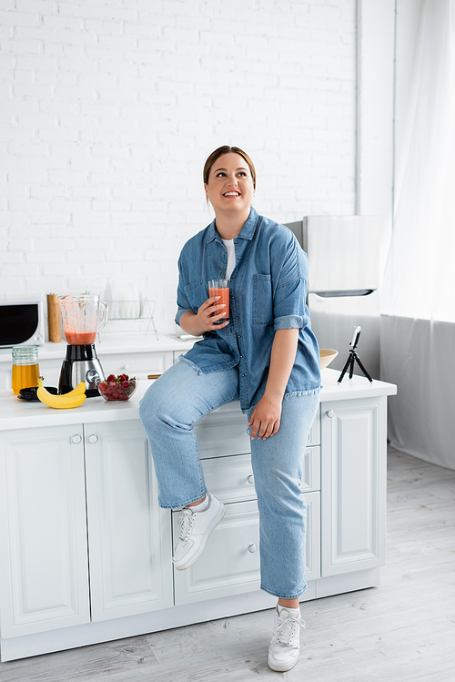 Happy plus size woman holding smoothie near fruits, blender and smartphone in kitchen