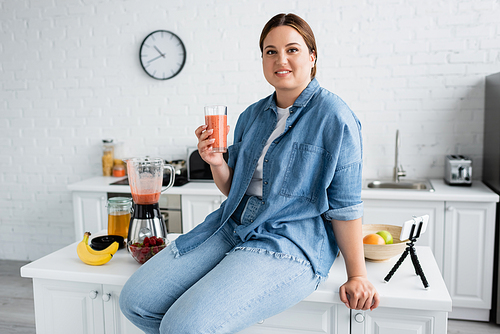 Smiling plus size woman holding glass of smoothie near fruits and smartphone on kitchen table