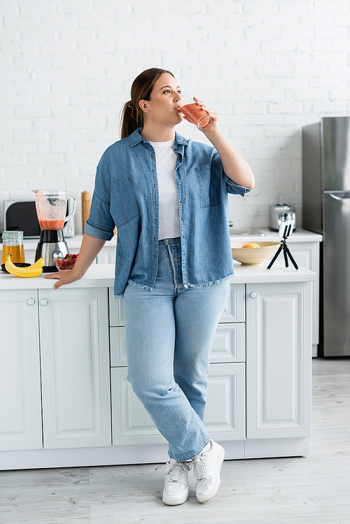 Young woman with overweight drinking smoothie near cellphone and ripe fruits in kitchen