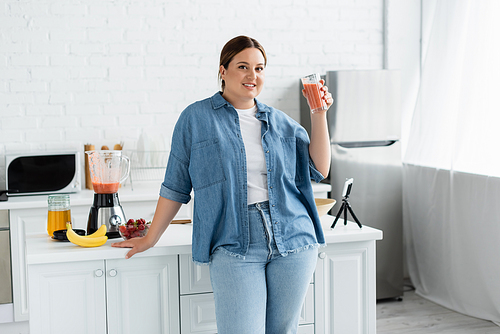 Smiling plus size woman holding glass of smoothie and  near cellphone in kitchen
