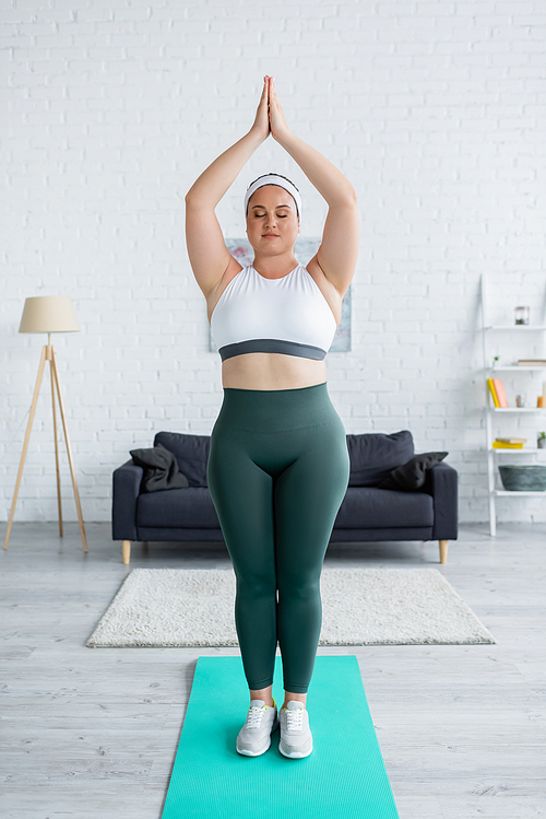 Plus size woman in sportswear standing with praying hands on fitness mat